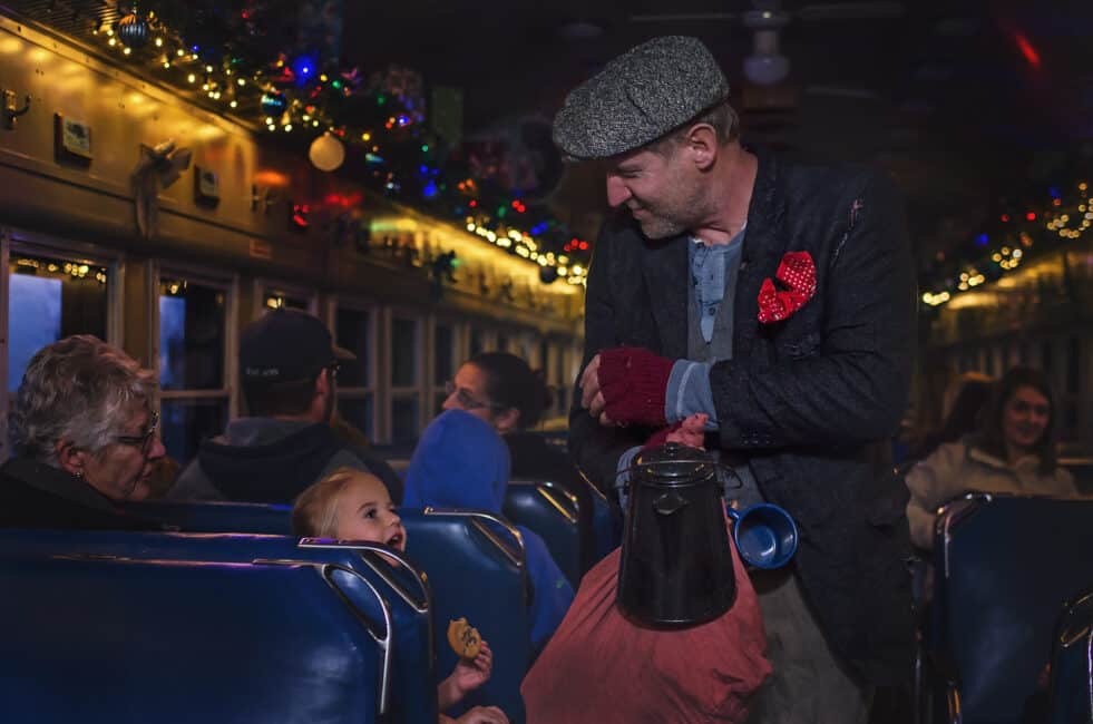 1. A man dressed in vintage attire with a flat cap and red gloves shares a moment with a young girl on a decorated train ride during the holidays.