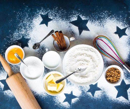 Fresh baking ingredients including flour, sugar, eggs, butter, cinnamon sticks, and cookies arranged on a star-themed surface with a rolling pin and colorful whisk.